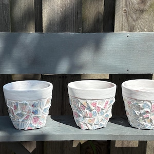 Puede incluir: Tres macetas de cerámica blanca con diseños de mosaicos. Los mosaicos son en tonos rosa, azul y blanco. Las macetas están sobre un banco gris frente a una valla de madera.