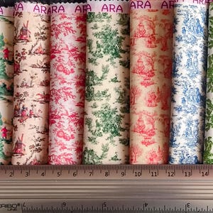 Op de afbeelding: Een verzameling opgerolde stofstalen met verschillende toile-patronen. De stoffen tonen scènes in rood, groen en blauw, met een liniaal die de afmetingen in centimeters aangeeft. Het woord "ARA" staat op de bovenrand van de stof.