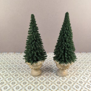 Könnte beinhalten: Zwei künstliche Mini-Weihnachtsbäume in weißen Töpfen vor einem grün-braunen gemusterten Hintergrund.