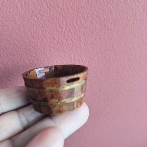 Puede incluir: Un pequeño cubo de madera con un acabado marrón y dorado. El cubo está hecho de duelas de madera y tiene un asa de metal.