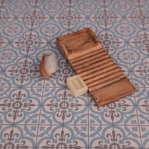 Puede incluir: Un juego en miniatura de tabla de lavar, jabón y cepillo de madera. La tabla de lavar es marrón con ranuras horizontales. Una pequeña pastilla de jabón con la palabra "SOAP" está al lado de la tabla de lavar. También hay un cepillo pequeño con mango de madera y cerdas blancas.