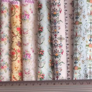 Puede incluir: Una colección de rollos de tela con estampados florales en varios colores, incluyendo rosa, amarillo y azul. Los diseños de tela presentan rosas, pájaros y adornos ornamentales. Los rollos están dispuestos verticalmente, con una regla en centímetros.