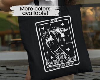 May include: Black tote bag with a white design of a tarot card. The card is labeled "The Tattoo Artist" and features a hand holding a tattoo machine. The card number is LXXVIII.