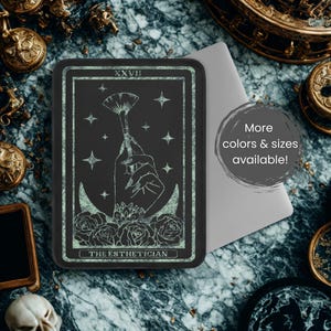 Op de afbeelding: Een zwart rechthoekig tarotkaartontwerp met een hand die een make-upkwast vasthoudt, omringd door sterren, rozen en een maan. De tekst "THE ESTHETICIAN" staat onderaan. De tekst "More colors & sizes available!" staat in een grijze cirkel.