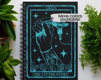 May include: Black spiral-bound notebook with a teal illustration of a hand holding a tattoo machine. The design includes stars, roses, and the text "THE TATTOO ARTIST" and "LXVII". A banner reads "More colors available!"