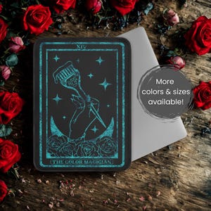 Può includere: Carta dei tarocchi rettangolare nera con dettagli turchese, raffigurante una mano che tiene un pennello e un coltello, con stelle e rose. Il testo recita "THE COLOR MAGICIAN". La carta è su una superficie di legno, circondata da rose rosse.