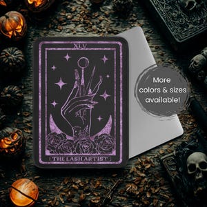 May include: A black rectangular phone case featuring a tarot card design with a hand holding lash tools. The design includes purple accents, stars, roses, and the text "THE LASH ARTIST" at the bottom. The text "XLV" is at the top.