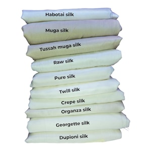 May include: A vertical stack of ten folded fabric swatches in shades of off-white and cream. Each layer is labelled with black text: Habotai silk, Muga silk, Tussah muga silk, Raw silk, Pure silk, Twill silk, Crepe silk, Organza silk, Georgette silk, Dupioni silk.
