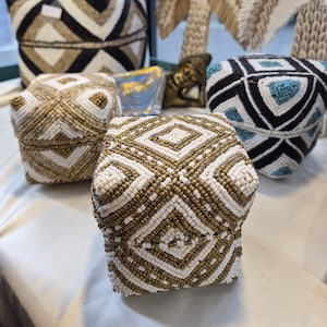 May include: A collection of square lidded boxes covered in intricate beadwork. The boxes feature geometric patterns in combinations of white, gold, black, and turquoise beads. Some boxes are stacked, while others sit individually on a flat surface.