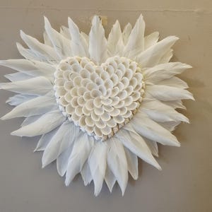 Puede incluir: Decoración mural en forma de corazón, hecha con conchas marinas y rodeada de plumas blancas. Las plumas están dispuestas en un patrón radial, creando un efecto floral. El fondo es de color gris claro neutro, resaltando los elementos blancos.