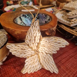 以下が含まれることがあります： 小さな白い貝殻で作られた、手作りの星型のオーナメント。星は吊り下げ用の天然繊維のコードに取り付けられています。背景には、他の貝殻を使った工芸品や木製のボウルがあります。