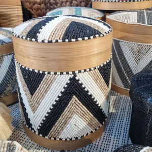 Op de afbeelding: Een ronde houten opbergcontainer met een deksel, versierd met een geometrisch patroon van zwarte, witte en beige kralen. De container heeft een natuurlijke houten afwerking en maakt deel uit van een collectie van vergelijkbare handgemaakte items.