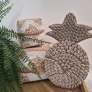Pode incluir: Um adorno de parede em forma de abacaxi coberto com pequenas conchas brancas, ao lado de duas caixas quadradas trançadas com um padrão geométrico branco e bege. Os itens estão dispostos juntos em uma bandeja de madeira clara.