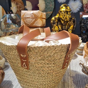 Könnte beinhalten: Eine gewebte Stroh-Tragetasche mit braunen Ledergriffen und -besatz. Die Tasche hat ein weißes Innenfutter und wird auf einer gemusterten Oberfläche präsentiert. Dekorative Fächer und eine goldene Gorillastatue sind im Hintergrund zu sehen.