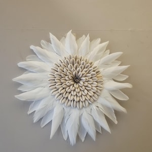 Puede incluir: Decoración mural con forma de flor. Los pétalos son de plumas blancas, y el centro está compuesto por pequeñas conchas marinas beige. La pieza está montada sobre una pared de color neutro, creando un contraste llamativo.