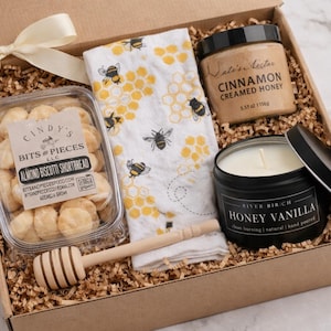 May include: A gift box with a cream bow, containing almond biscotti, a bee-themed tea towel, cinnamon creamed honey, a honey vanilla candle, and a wooden honey dipper. The box is filled with shredded paper.