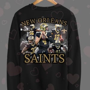 Peut inclure: Sweat-shirt noir avec un graphique de joueurs de football des Saints de la Nouvelle-Orléans. Le texte "NEW ORLEANS SAINTS" est imprimé en or au dos du sweat-shirt.