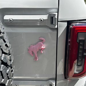 May include: A light pink, three-dimensional emblem of a bucking horse mounted on the white rear panel of a vehicle. The emblem is positioned between a large black tire on the left and a vertical red and clear taillight assembly on the right.