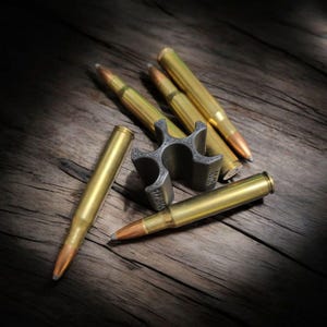 May include: A collection of brass-colored rifle cartridges and a silver-colored bottle opener with a unique, multi-pronged design. The cartridges have copper-colored tips and are arranged on a weathered wooden surface, creating a rustic aesthetic.