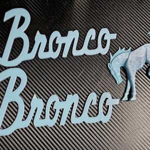 May include: Two light blue plastic emblems featuring the word Bronco in a cursive script font, accompanied by a separate light blue plastic emblem of a bucking horse. The surfaces of these three-dimensional items show a layered texture.