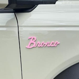 May include: A light cream-colored vehicle exterior panel featuring a script-style emblem in a matte pink finish. The emblem displays the word Bronco in a cursive font, positioned horizontally on the side of the vehicle near the front fender.