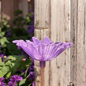 May include: A lavender-colored, flower-shaped garden ornament. The flower has textured petals and is mounted on a light brown stake. The background includes wooden planks and purple flowers.