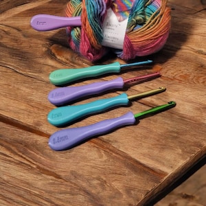 May include: A collection of crochet hooks with colorful handles in shades of purple, teal, and green, alongside a ball of multicolored yarn. The hooks are labeled with sizes, including 4mm, 5.5mm, and 6mm. The yarn has a label that reads "Facets".