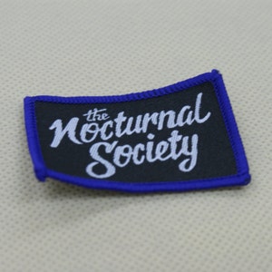 Puede incluir: Un parche bordado negro y azul con texto blanco que dice "the Nocturnal Society".