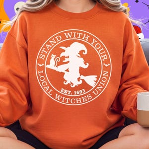 May include: An orange crewneck sweatshirt featuring a white circular graphic on the chest. The graphic depicts a silhouette of a witch on a broomstick with a cat and the text "STAND WITH YOUR LOCAL WITCHES UNION EST. 1693" in a circular border.