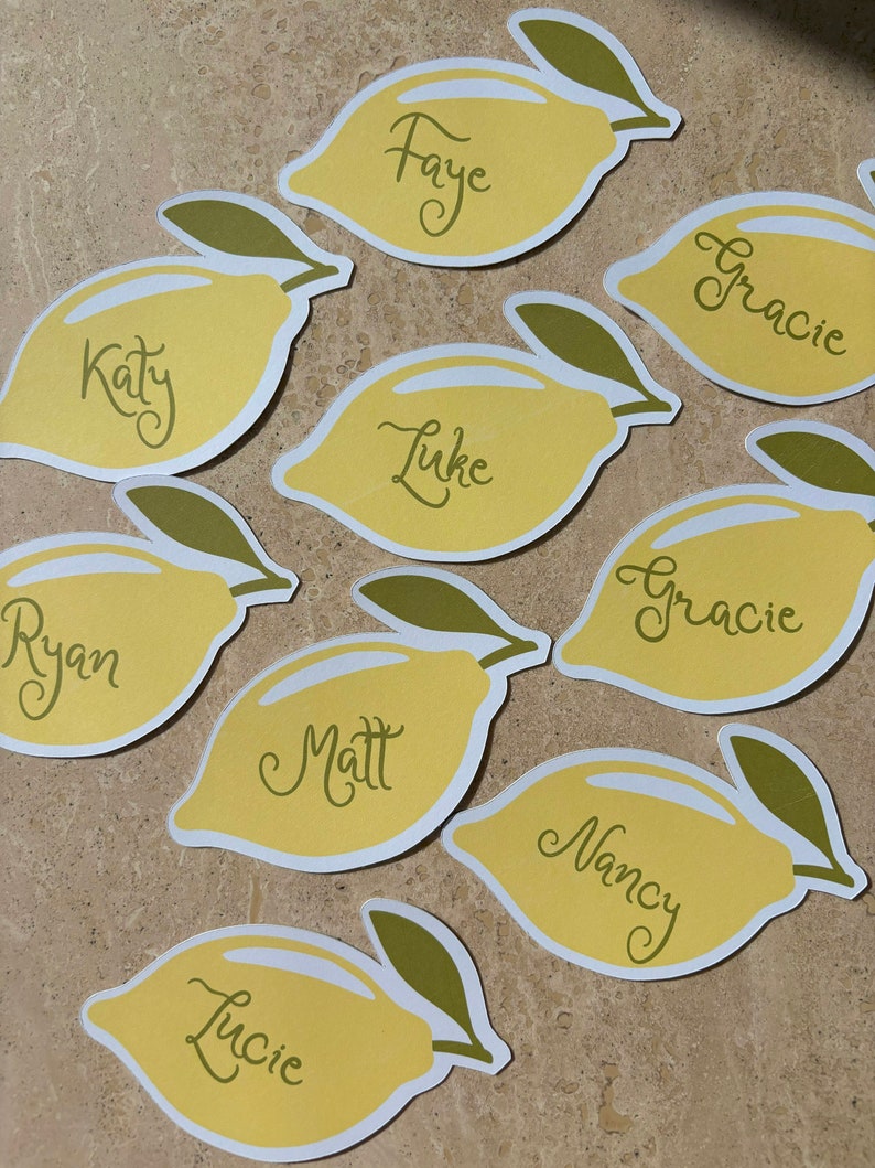 May include: Nine yellow lemon-shaped place cards with green stems and leaves. The names Faye, Katy, Luke, Ryan, Matt, Gracie, Nancy, and Lucie are written on the cards.