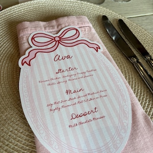 Op de afbeelding: Een roze en witte menukaart met een strik, met de naam "Ava" en een gedetailleerd menu. De kaart ligt op een roze servet, met bestek en een placemat op de achtergrond.