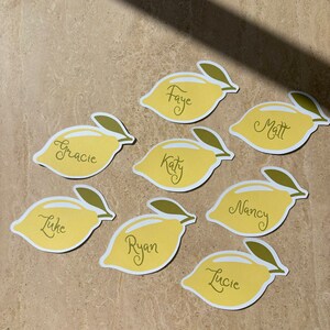 May include: Eight yellow lemon-shaped place cards with green leaves, each with a name written in black cursive script: Faye, Matt, Gracie, Katy, Luke, Ryan, Nancy, and Lucie.