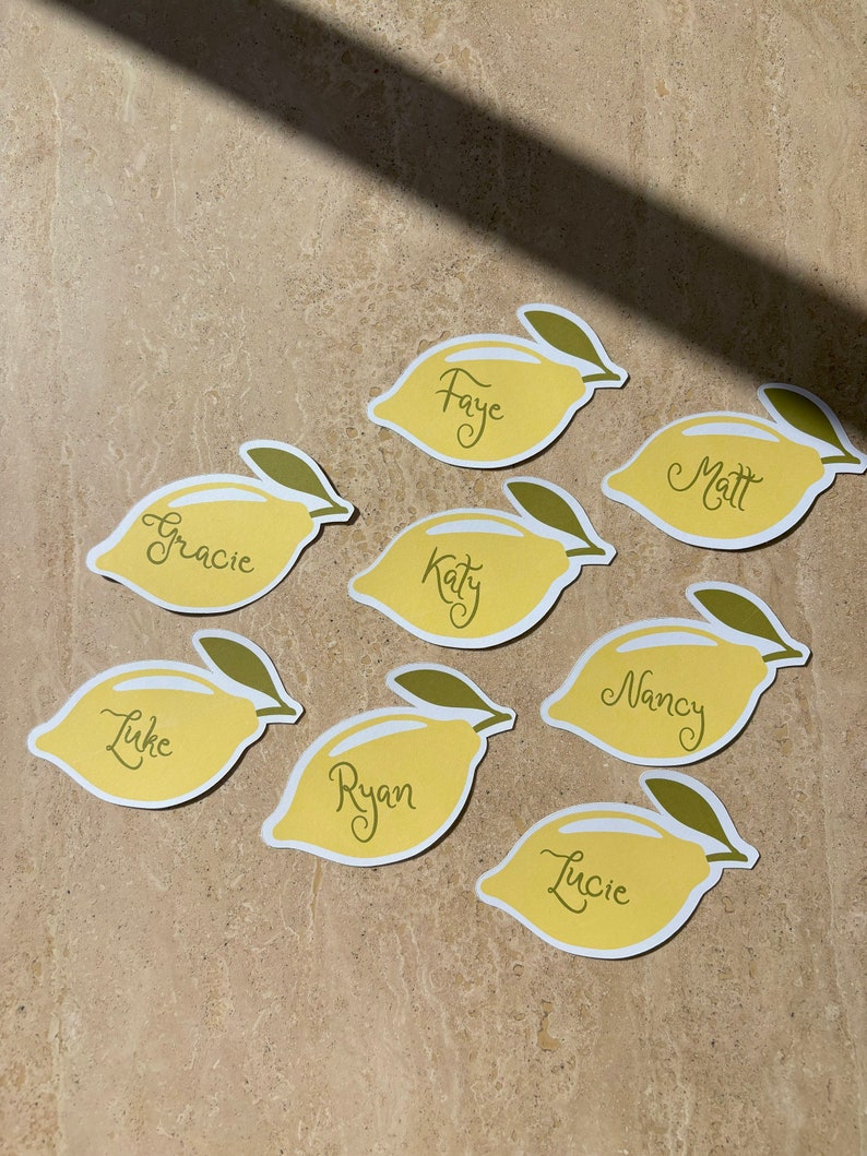 May include: Eight yellow lemon-shaped place cards with green leaves, each with a name written in black cursive script: Faye, Matt, Gracie, Katy, Luke, Ryan, Nancy, and Lucie.