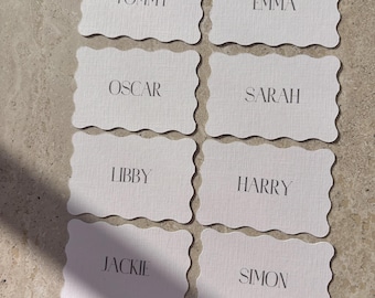 Könnte beinhalten: Sechs weiße Platzkarte mit gewellten Rändern und schwarzem Text. Die Namen auf den Karten sind Tommy, Emma, Oscar, Sarah, Libby, Harry, Jackie und Simon.