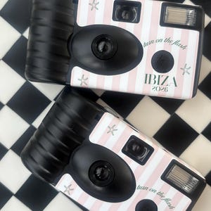 Peut inclure: Deux appareils photo jetables noirs et roses avec le texte "IBIZA 2026" et "turn on the flash". Les appareils photo ont un motif à rayures roses et blanches et des illustrations d'étoiles de mer. Sur une surface à carreaux noirs et blancs.