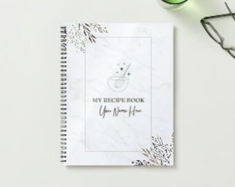 May include: A white spiral bound recipe book with a marble pattern and gold foil accents. The cover features a stylized illustration of a mixing bowl with a whisk and stars. The text "MY RECIPE BOOK" and "Your Name Here" is printed in black.