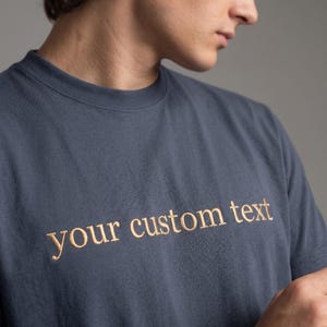 Może przedstawiać: Szara koszulka z haftowanym złotym napisem "your custom text". Koszulka ma okrągły dekolt i wydaje się być wykonana z miękkiego materiału. Zdjęcie przedstawia osobę noszącą koszulkę.