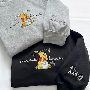 May include: Three sweatshirts, gray and black, with embroidered Winnie the Pooh designs. The gray sweatshirts say "baba bear" and "Aminah" on the sleeve. The black sweatshirt says "mama bear" with a Winnie the Pooh design.