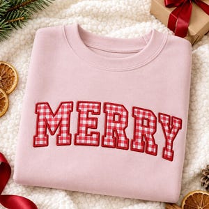 Op de afbeelding: Een lichtroze sweater met ronde hals en het woord MERRY in grote blokletters. De letters zijn gevuld met een rood-wit vichy-ruitpatroon en omlijnd met rood borduurgaren, wat zorgt voor een gestructureerde, op universiteitsstijl geïnspireerde look.