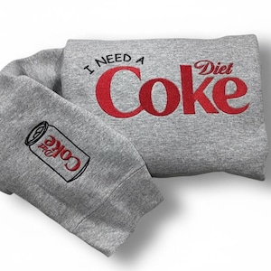 Op de afbeelding: Een grijze sweatshirt met de tekst "I NEED A Diet Coke" in het rood. De mouw heeft een geborduurde Diet Coke-blik. De sweatshirt is op een witte achtergrond gevouwen.
