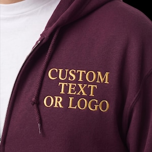 May include: A burgundy zip-up hoodie with the embroidered text "CUSTOM TEXT OR LOGO" in gold. The hoodie features a drawstring hood and a white undershirt is visible beneath the zipper.