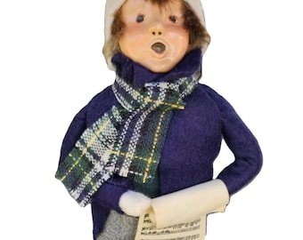 May include: A figurine of a child dressed for winter, wearing a blue sweater, gray pants, and a white hat. The child is holding a scroll of sheet music and has a green base.