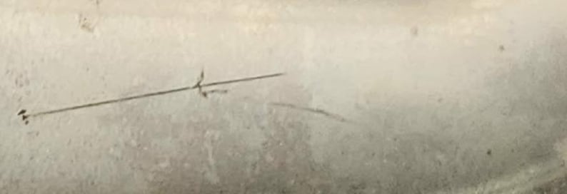 May include: A close-up of a light-colored, textured surface with a thin, straight black line and a small, stylized arrow pointing to the left. There is a curved black line below the straight line.