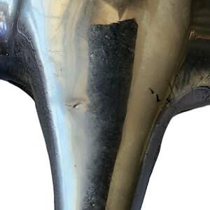 May include: Close-up of a tarnished, metallic object with a curved, flared shape. The surface reflects light, showing imperfections and wear. The object appears to be a decorative or functional piece, possibly part of a larger item.