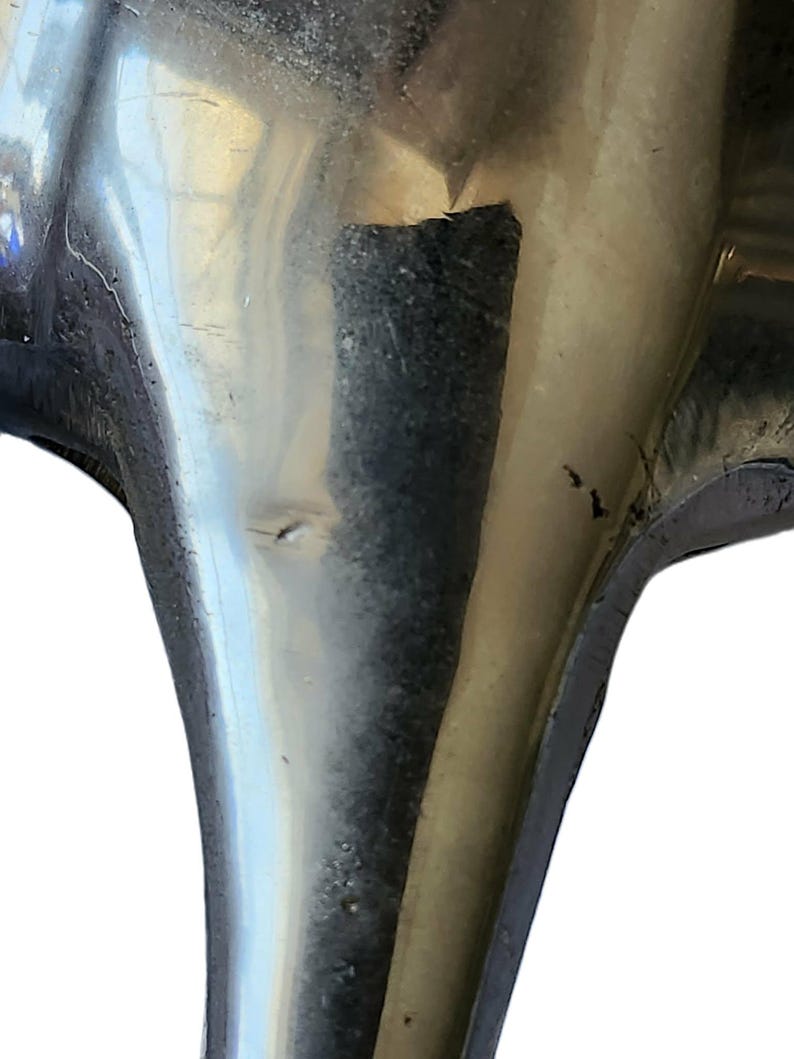 May include: Close-up of a tarnished, metallic object with a curved, flared shape. The surface reflects light, showing imperfections and wear. The object appears to be a decorative or functional piece, possibly part of a larger item.