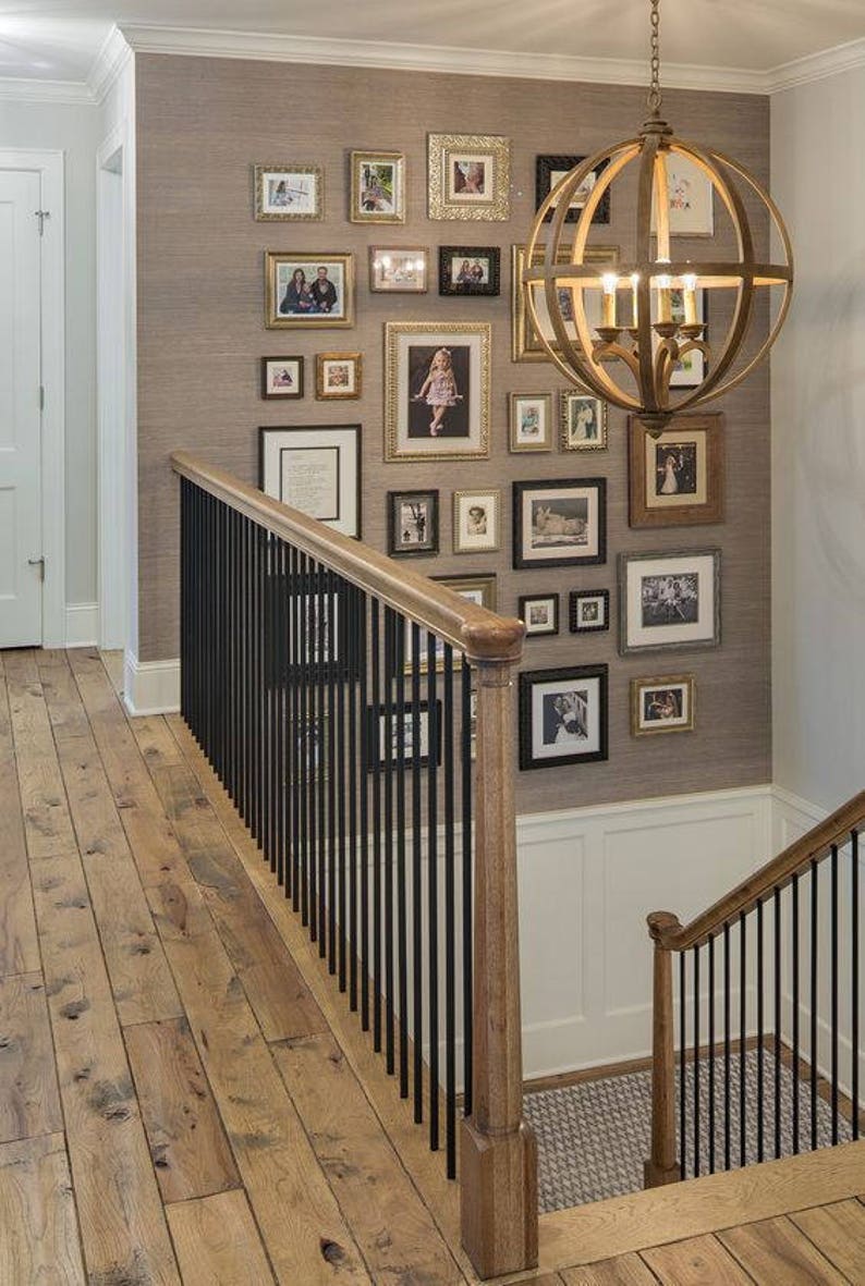 Great example of an asymmetrical, gallery-style group of photos on a wall encompassing the landing on a set of stairs as well as the second floor. Various sizes of black and gold frames with white mats on a light brown wall.