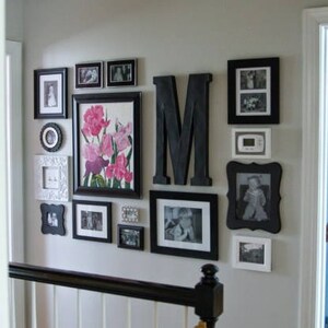 Strong example of an asymmetrical, mostly monochromatic, gallery-style display of black and white framed photos mixed with an oversized black monogram. Frames have either white mats or no mats. One large, colorful floral print with pink and purple.