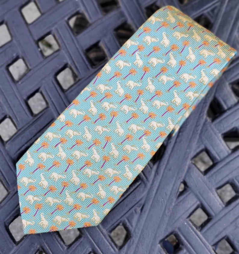 May include: A patterned necktie featuring a repeating design of white elephants and orange palm trees on a light blue background. The tie is made of a textured fabric and is displayed on a dark blue surface.