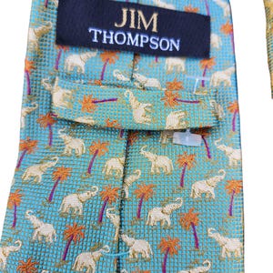 May include: A close-up of a Jim Thompson necktie. The tie features a repeating pattern of white elephants and orange palm trees on a turquoise background. The Jim Thompson label is visible in navy blue with white text.