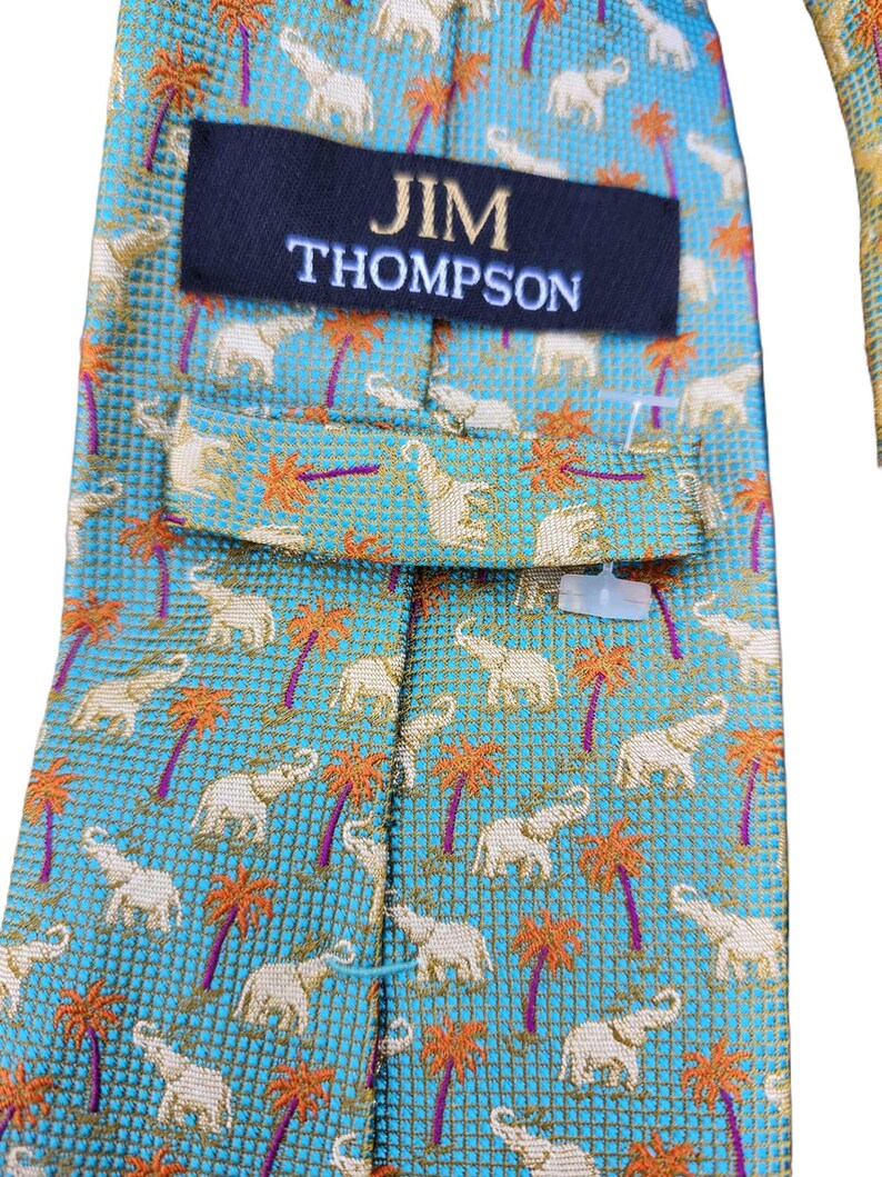 May include: A close-up of a Jim Thompson necktie. The tie features a repeating pattern of white elephants and orange palm trees on a turquoise background. The Jim Thompson label is visible in navy blue with white text.